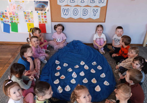 Dzieci z grupy Stokrotki siedzą wokół dużej kropli wody i losują małe krople opowiadając do czego służy woda.