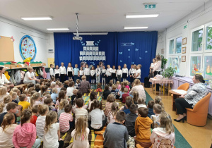 Dzieci ubrane na galowo stoją na tle dekoracji, przed nimi siedzą pozostałe dzieci oraz panie.