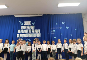 Dzieci stoją na tle dekoracji, jedna dziewczynka trzyma w ręku mikrofon.