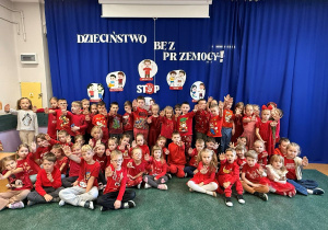 Dzieci z grup sześciolatków, ubrane w czerwone koszulki, pozują do zdjęcia na tle informacji o kampanii Dzieciństwie Bez Przemocy.