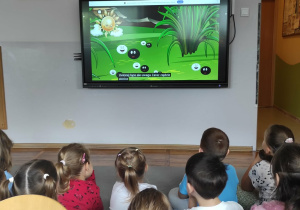 Dzieci z grupy Misie siedzą na dywanie oglądając film edukacyjny wyświetlany na tablicy multimedialnej dotyczący Dnia Kropki.
