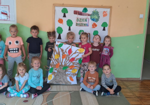 Dzieci z grupy „Misie” pozują do zdjęcia na tle tablicy dekoracyjnej w rękach trzymają pracę plastyczną „Drzewko”, które wykonały z szarego papieru na dużym białym brystolu i przykleiły liście ze swoimi imionami.