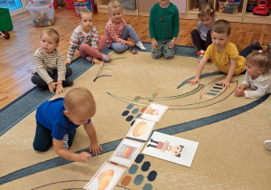 Dzieci z grupy Biedronki siedzą na dywanie, chłopiec układa historyjkę obrazkową dotyczącą wypiekania chleba.