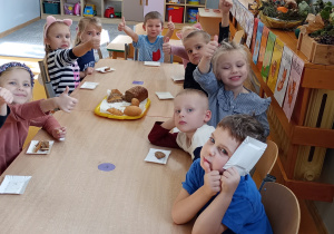 Dzieci siedzą dookoła stolika z pieczywem i pokazują kciukiem ok.