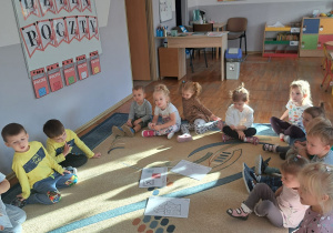 Dzieci siedzą w kole na dywanie, pośrodku są rozłożone plansze związane z pocztą.