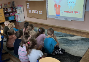 Dzieci siedzą na dywanie i oglądają film na tablicy multimedialnej.