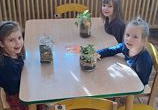 Dziewczynki siedzą przy stoliku prezentując swoje kompozycje „Las w słoiku”.