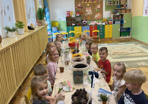 Dzieci siedzą przy stolikach na którym stoją słoiki oraz materiały przyrodnicze: ziemia, piasek, kamyczki, szyszki, kasztany, żołędzie oraz roślinki.