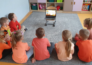 Dzieci siedzą na dywanie i oglądają film edukacyjny na laptopie.