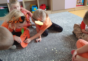 Dzieci siedząc na dywanie karmią papierową dynię pestkami z cyferkami.