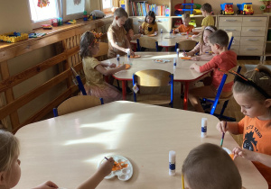 Dzieci siedzą przy stoliczkach i malują farbami talerzyki papierowe, z których później wykonają dynię.