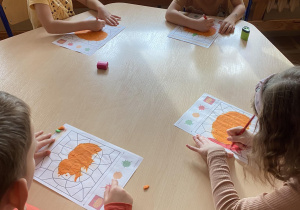 Dzieci siedzą przy stoliczku i wykonują pracę plastyczną: wylepiają dynię pomarańczową plasteliną, a następnie kolorują tło na czerwono.