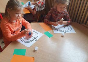 Dziewczynki wykonują prace plastyczną wyklejając dynie pomarańczowym i zielonym papierem.