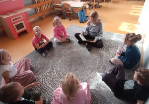 Oliwia czyta książkę dzieciom, które siedzą w kręgu na dywanie.