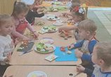 Dzieci siedzą przy stolikach mają przed sobą deski do krojenia oraz nożyki. Na stolikach stoją talerze z warzywami i pieczywem.