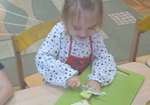 Dziewczynka siedzi przy stoliku na desce do krojenia leży chleb posmarowany masłem i kroi ogórka.