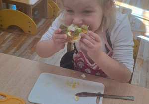 Dziewczynka siedzi przy stoliku i zjada kanapkę zrobioną przez siebie.
