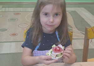 Dziewczynka siedzi przy stoliku i trzyma w rękach kanapkę zrobioną przez siebie.
