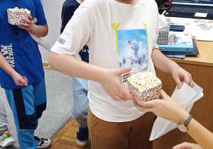 Chłopiec w białej bluzce odbiera od nauczycielki kubeczek z popcornem.