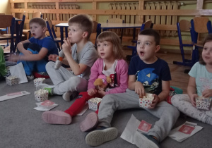 Dzieci siedzą na dywanie, oglądają bajkę oraz zajadają popcorn.