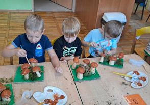 Dzieci siedzą przy stoliku i malują brązową farbą swoje grzybki.