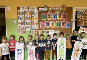 Dzieci stoją przed dekoracją z okazji Dnia Kredki i trzymają w rękach pokolorowane przez siebie w zespołach ogromne kredki