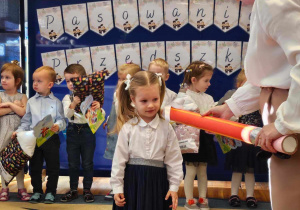 Pani Dyrektor kładąc na lewym ramieniu dużą kredkę, pasuje dziewczynkę z grupy „Biedronki” na przedszkolaka.