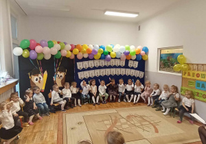 Dzieci siedzą na krzesełkach na tle dekoracji „Pasowanie na Przedszkolaka”.