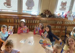 Dzieci siedzą przy stoliku i przedstawiają swoje prace.