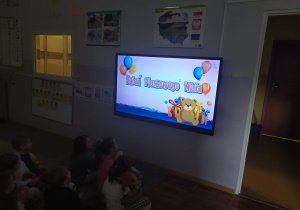 Dzieci siedzą na dywanie i oglądają film edukacyjny na tablicy multimedialnej o historii pluszowego misia.
