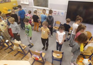 Dzieci sadzają misie na krzesełkach zgodnie z instrukcją nauczyciela: na, pod, obok.