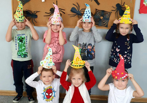 Dzieci z grupy Liski pozują do wspólnego zdjęcia, prezentując wykonane przez siebie kapelusze andrzejkowe. W tle tablica z dekoracją andrzejkową.