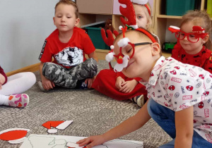 Dzieci siedzą na dywanie, wykonują zadanie uzupełniają planszę odpowiednim kształtem czapki Mikołaja.