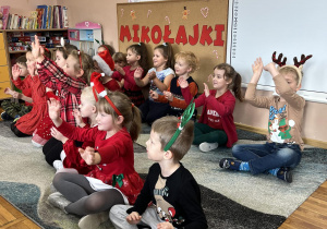 Dzieci siedzą na dywanie i śpiewają piosenkę dla Mikołaja.