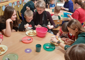 Dzieci wraz z rodzicami przy stoliku ozdabiają swoje smakołyki.