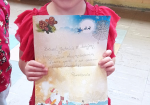 Zdjęcie przedstawia dziewczynkę w czerwonej sukience trzymającą trzeci list od św. Mikołaja znaleziony w przedszkolnej szatni
