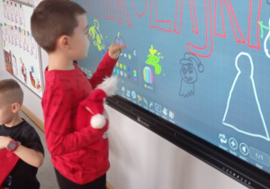 Zdjęcie przedstawia chłopca w czerwonej bluzce stojącego przy monitorze. Chłopiec rysuje mikołajkową zagadkę w grze kalambury dla reszty grupy.