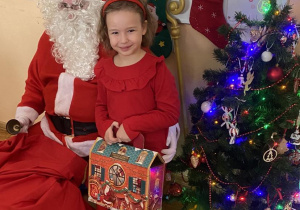 Dziewczynka w czerwonej sukience pozuje do zdjęcia z Mikołajem.