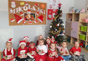 Dzieci z grupy Liski siedzą przed choinką, trzymają prezenty od Świętego Mikołaja.