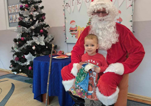 Chłopiec pozuje do zdjęcia z Mikołajem, trzyma otrzymany prezent.
