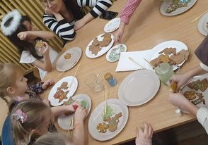 Dzieci siedzą przy stoliku i wraz z rodzicami dekorują pierniczki.