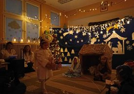Dzieci siedzą na dywanie przy szopce, dziewczynka przebrana za aniołka trzyma koszyczek i sypie gwiazdkami papierowymi.