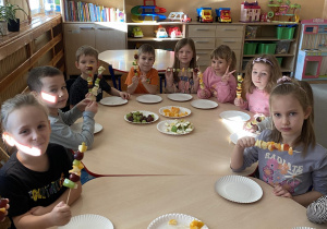 Dzieci siedzą przy stoliku i pokazują samodzielnie wykonane szaszłyki.