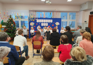 Dzieci stoją na tle dekoracji i recytują wiersze dla Babć i Dziadków siedzących na widowni.