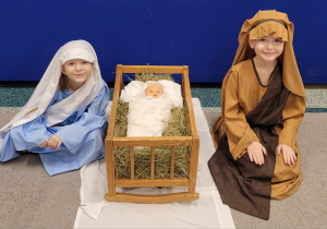 Dziewczynka i chłopiec w strojach Maryi i Józefa siedzą przy żłóbku.
