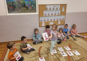 Dzieci siedzą na dywanie, wskazują obrazek wg opisu nauczyciela.