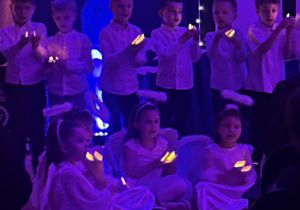 Zdjęcie przedstawia występ dzieci z grupy „Pszczółki”, dziewczynki w strojach aniołków siedzą na podłodze a chłopcy ubrani na galowo stoją z tyłu. Wszystkie dzieci trzymają w dłoniach zaświecone lampeczki