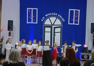 Zdjęcie przedstawia występ dzieci z grupy „Smerfy”, po lewej strony stoi dziewczyna w stroju aniołka; pięć dziewczynek w białych bluzkach, sukienkach w kwiatach i wiankach na głowie; dwóch króli; dwóch pastuszków.