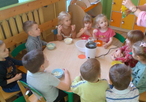 Dzieci siedzą przy stoliku i obserwują jak nauczyciel wsypuje ziarna kukurydzy do maszynki