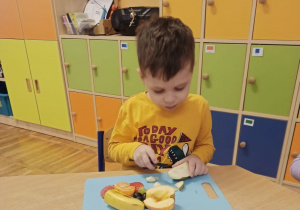 Chłopiec w żółtej bluzie siedzi przy stoliku i kroi na desce gruszkę plastikowym nożem.